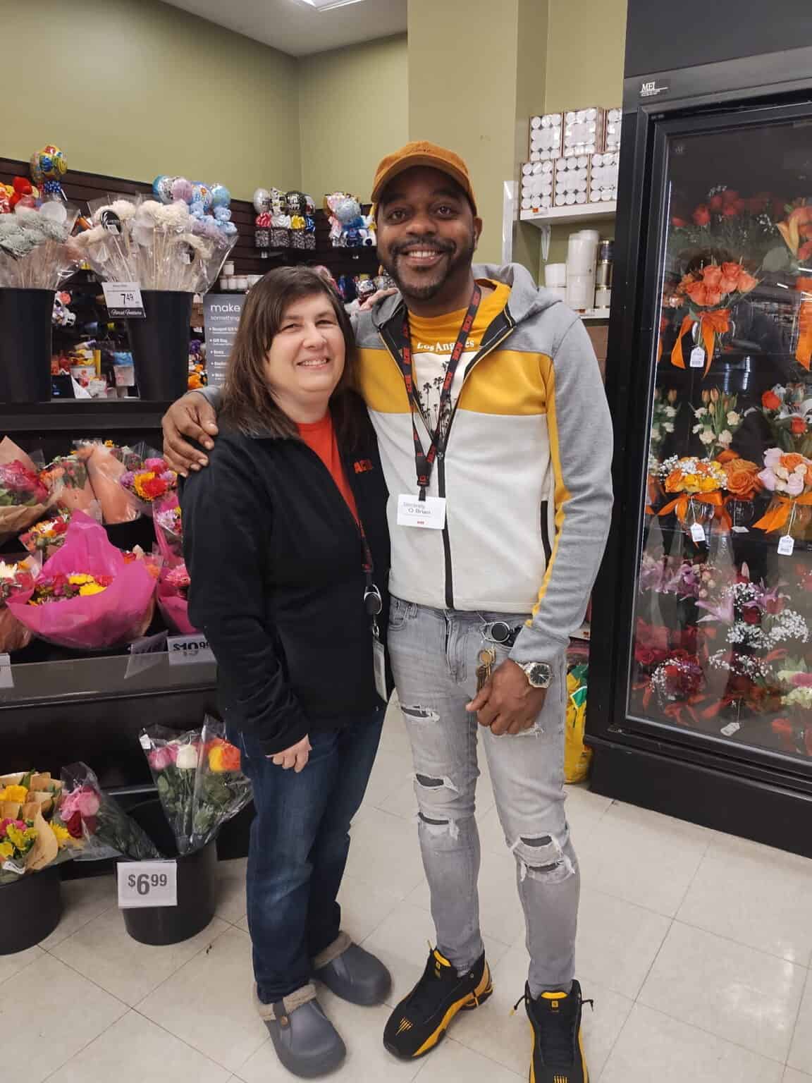 Smiling man and woman in a grocery store with flowers and products behind them.