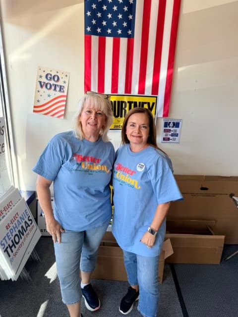 Two women wearing blue 'Better Union' T-shirts at a voting event with American flag and campaign ads.