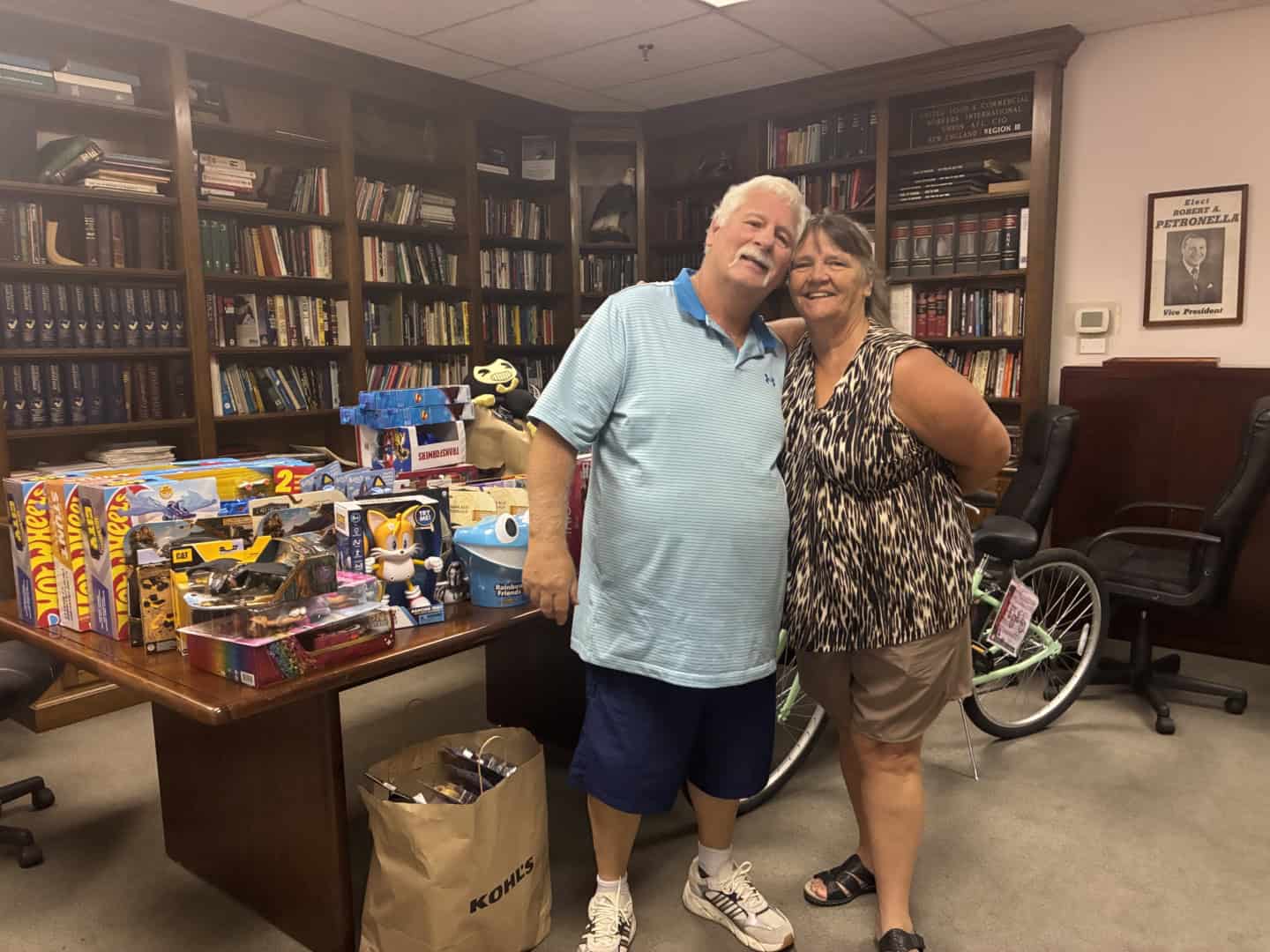 Two smiling people standing in a library with toys and books on a table behind them.