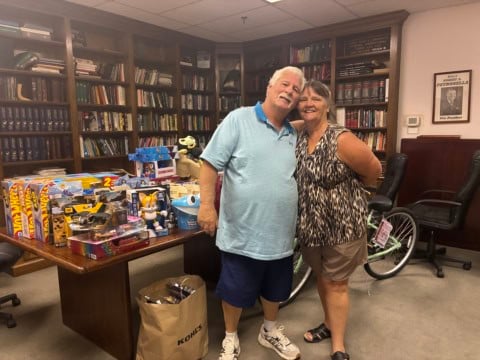 Two smiling people standing in a library with toys and books on a table behind them.