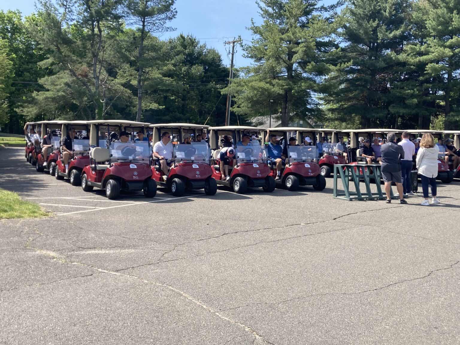 Group of people in golf carts parked in a lot with trees in the background.