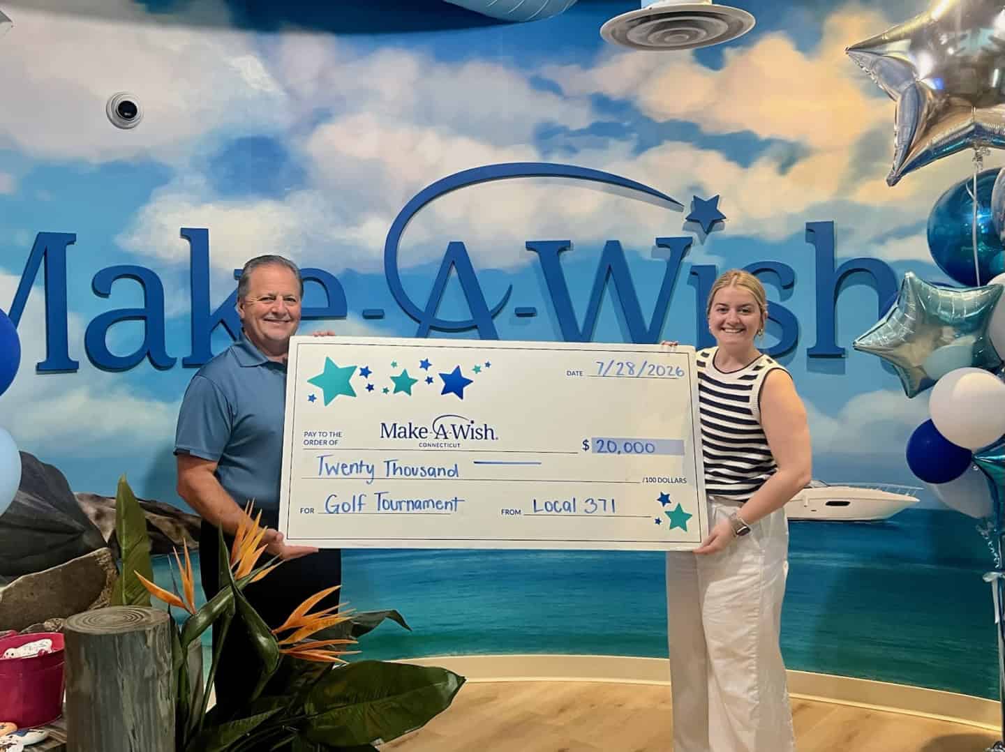 Two people holding a large check for $20,000 at a Make-A-Wish event with a beach-themed backdrop.