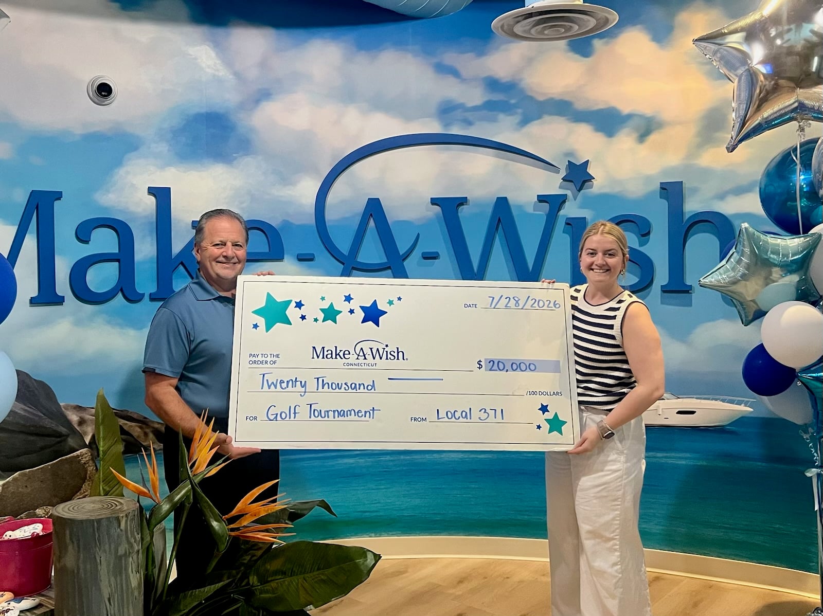 Two people holding a large check for $20,000 at a Make-A-Wish event with a beach-themed backdrop.