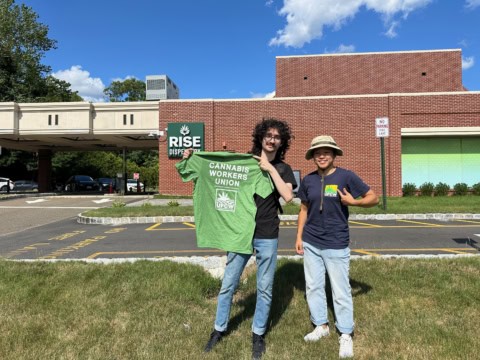 Two workers holding a Cannabis Workers Union shirt outside RISE Dispensary.